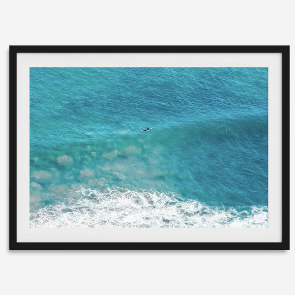 Aerial beach wall art featuring a lone surfer paddling through turquoise ocean waves, with foamy white surf along the shoreline.