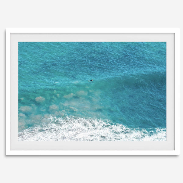 Aerial beach wall art featuring a lone surfer paddling through turquoise ocean waves, with foamy white surf along the shoreline.