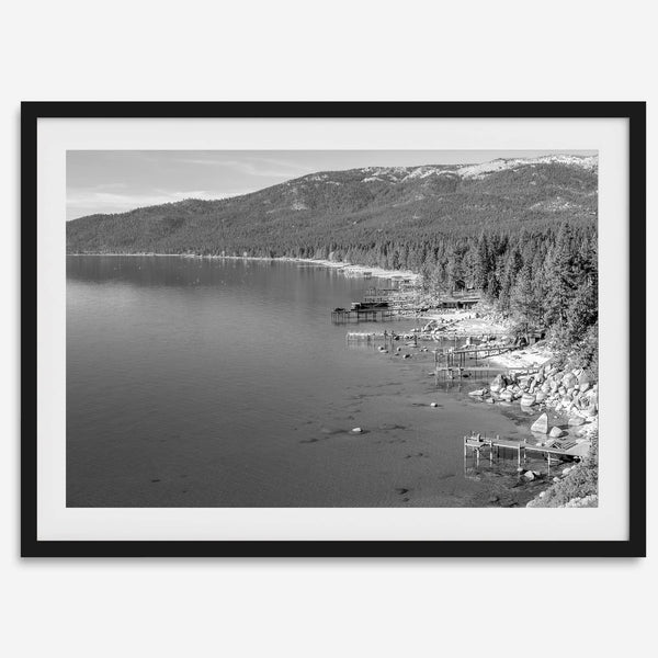 A fine art black and white Lake Tahoe print showcasing docks and boats nested on the lake waters on the left, while the beachfront, nestled against a backdrop of a lush forest and majestic mountain, paints a picturesque scene on the right.