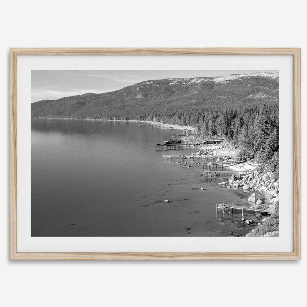 A fine art black and white Lake Tahoe print showcasing docks and boats nested on the lake waters on the left, while the beachfront, nestled against a backdrop of a lush forest and majestic mountain, paints a picturesque scene on the right.