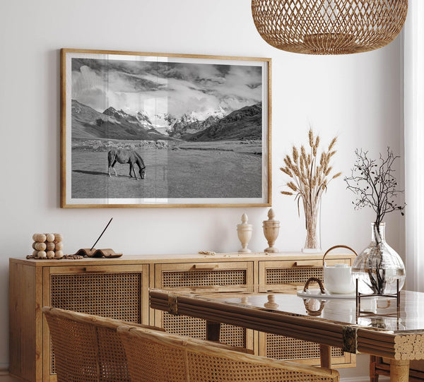 A black and white mountain horse fine art print that can come framed on unframed and was taken in Peru. This mountain wall art showcases snow-covered mountains in Peru and a charming horse grazing relaxingly in the forefront of the picture.