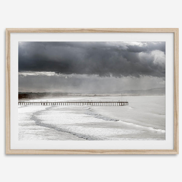 This fine art ocean photography print was taken in Pismo Beach, California, and showcases a distant view of an ocean pier and stormy clouds. Looking long and hard, you can see tens of surfers around the pier.