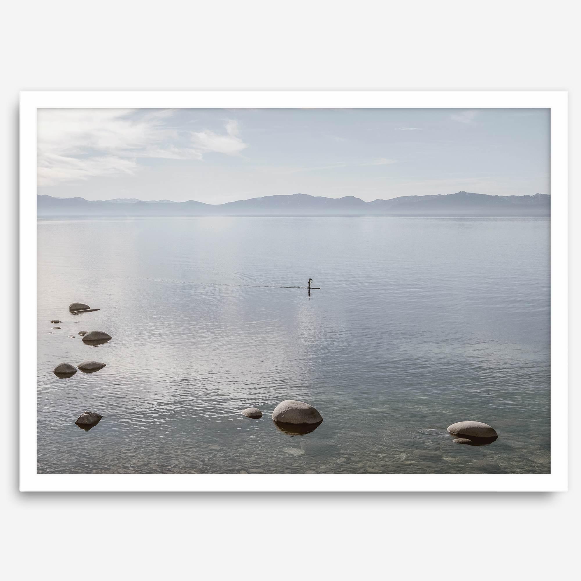 Fine art photography print of Lake Tahoe featuring a serene blue water surrounded by Pacific Northwest mountains, with a lone stand-up paddle silhouette on the lake.
