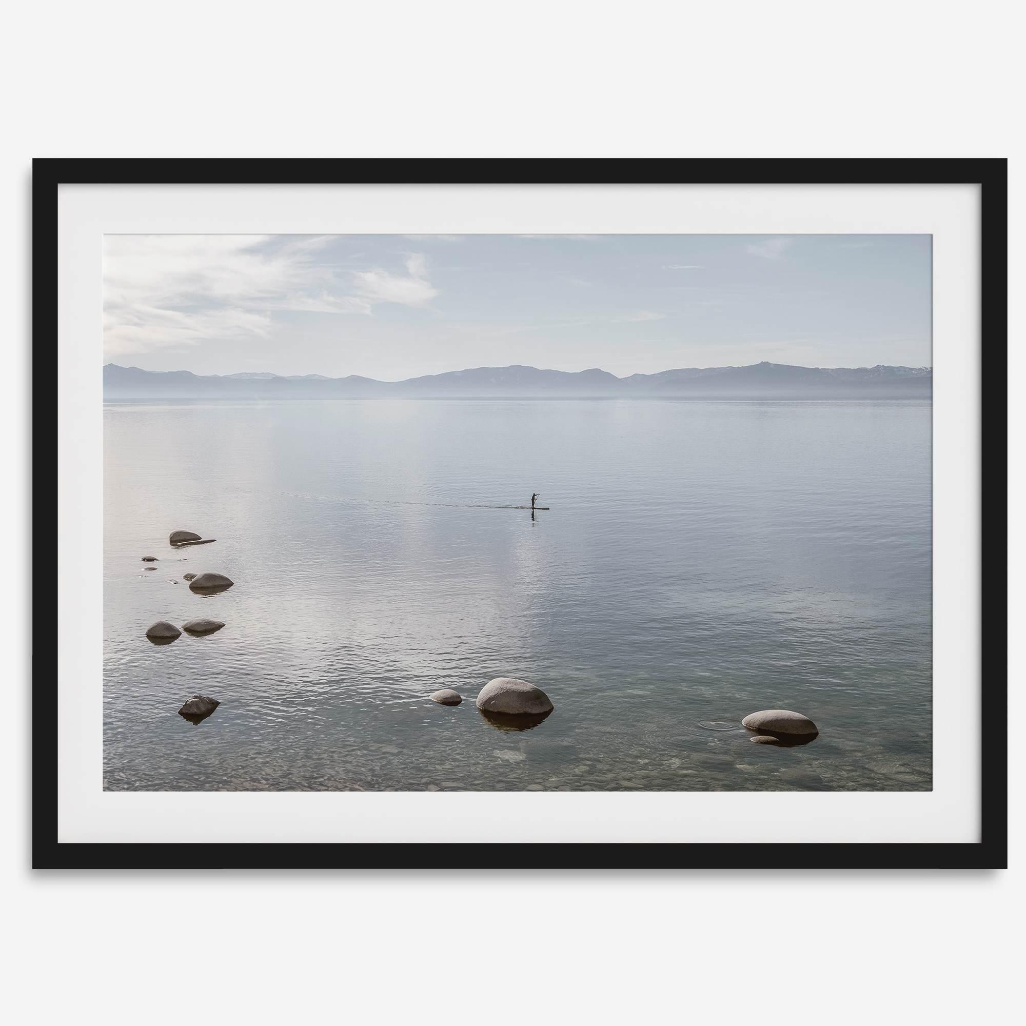 Fine art photography print of Lake Tahoe featuring a serene blue water surrounded by Pacific Northwest mountains, with a lone stand-up paddle silhouette on the lake.