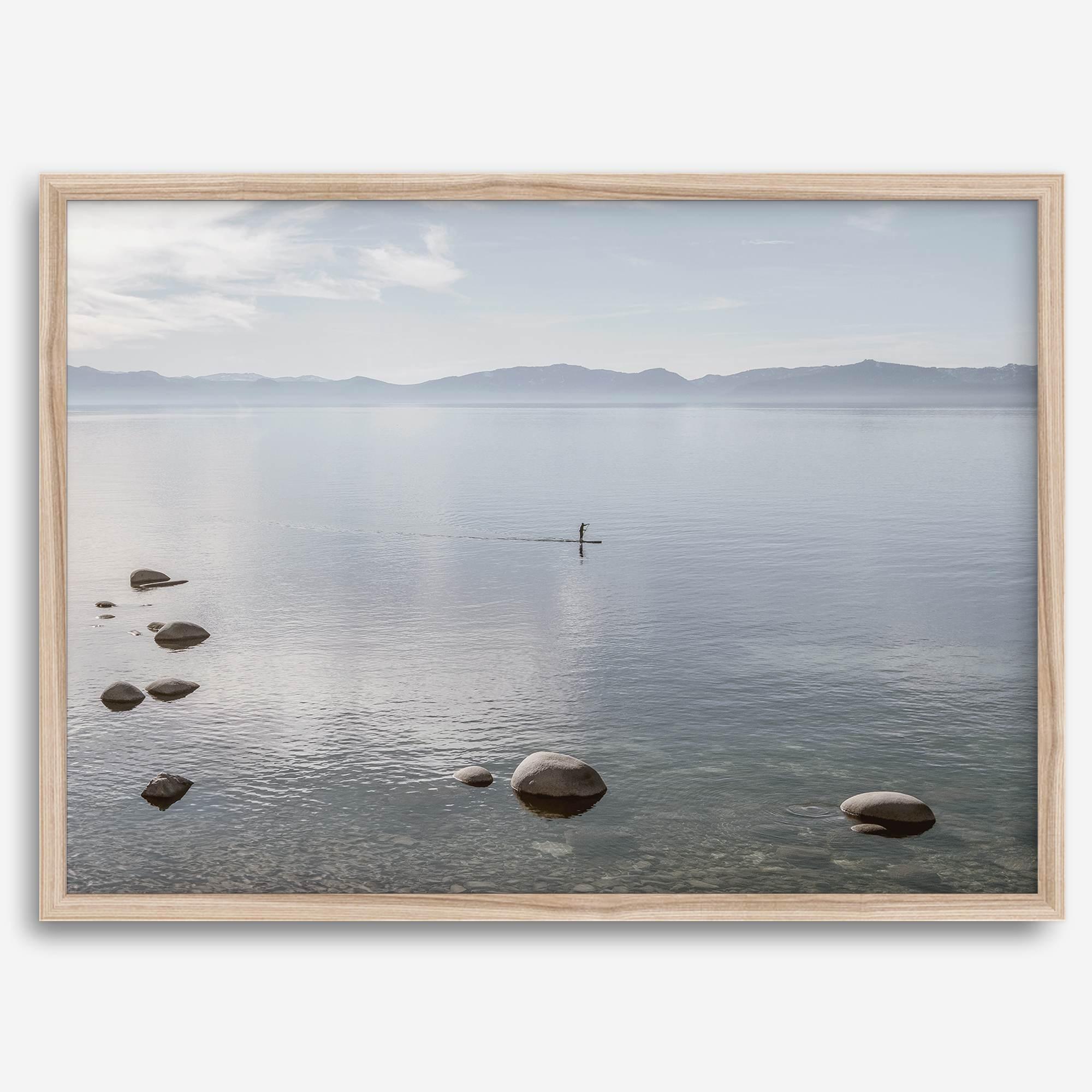 Fine art photography print of Lake Tahoe featuring a serene blue water surrounded by Pacific Northwest mountains, with a lone stand-up paddle silhouette on the lake.