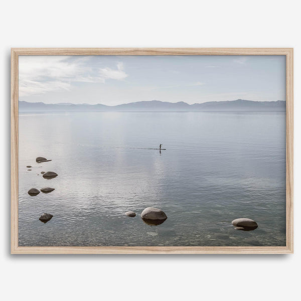 Fine art photography print of Lake Tahoe featuring a serene blue water surrounded by Pacific Northwest mountains, with a lone stand-up paddle silhouette on the lake.