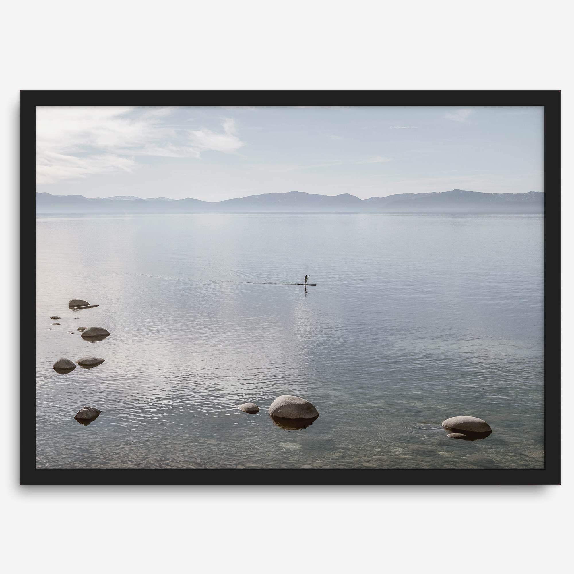 Fine art photography print of Lake Tahoe featuring a serene blue water surrounded by Pacific Northwest mountains, with a lone stand-up paddle silhouette on the lake.