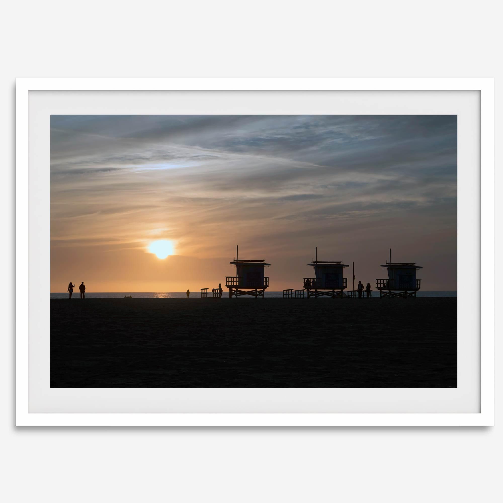 Step into Venice Beach with our fine art beach sunset framed or unframed print. The print showcases the beach in sunset with the silhouettes of the famous lifeguard towers and people in the sunset.
