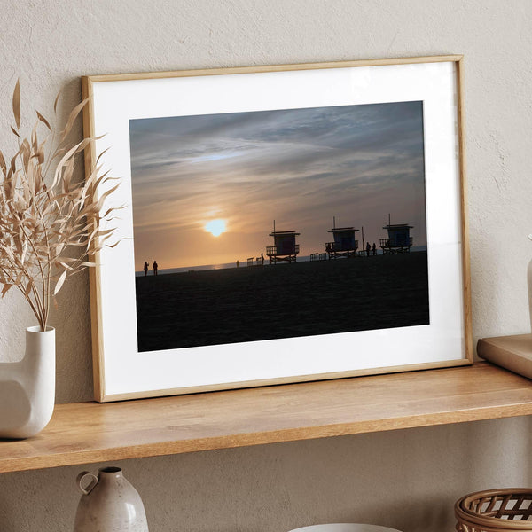 Step into Venice Beach with our fine art beach sunset framed or unframed print. The print showcases the beach in sunset with the silhouettes of the famous lifeguard towers and people in the sunset.