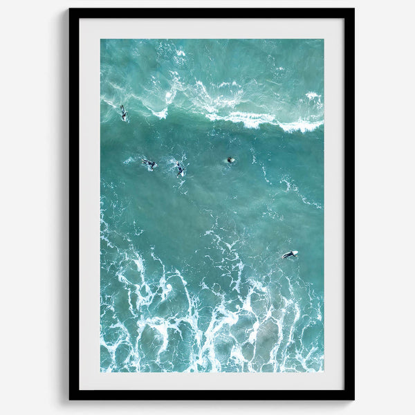 Vertical aerial photo of surfers paddling out to catch a wave at sunset, with wave trails visible in the water.