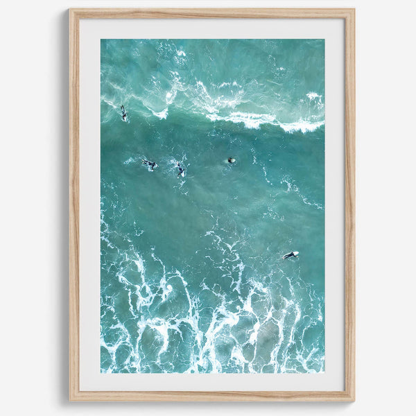 Vertical aerial photo of surfers paddling out to catch a wave at sunset, with wave trails visible in the water.