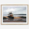 Weathered shipwreck resting on a quiet coastal shore, surrounded by marshland and a moody, overcast sky.