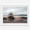 Weathered shipwreck resting on a quiet coastal shore, surrounded by marshland and a moody, overcast sky.