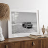 Black and white vintage beach print featuring a classic car parked at the edge of the ocean, capturing a retro coastal scene with timeless charm.