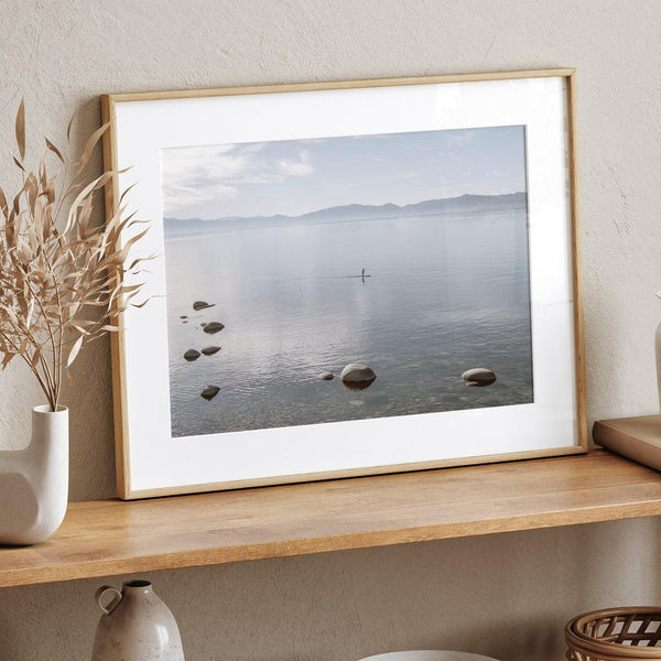 Fine art photography print of Lake Tahoe featuring a serene blue water surrounded by Pacific Northwest mountains, with a lone stand-up paddle silhouette on the lake.