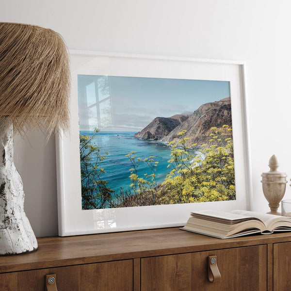 Capture the awe-inspiring coastal beauty with our fine art coastal print. Bixby Bridge spans the cliffs amidst vibrant yellow flowers, complementing the serene blue ocean. Perfect for framed or unframed wall art display.