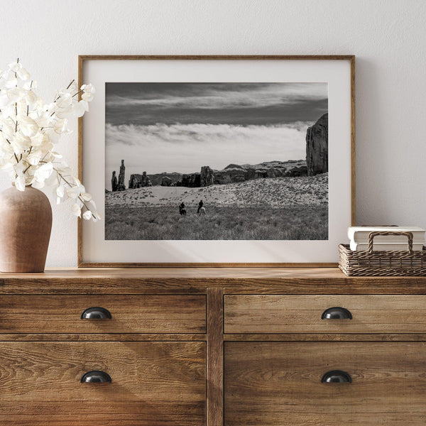 This Utah desert fine art photography print features two horse riders riding toward the horizon of the Utah desert with the unique towering rocks of Monument Valley as the backdrop. The dramatic cloudy sky adds intensity and beauty to the scene.