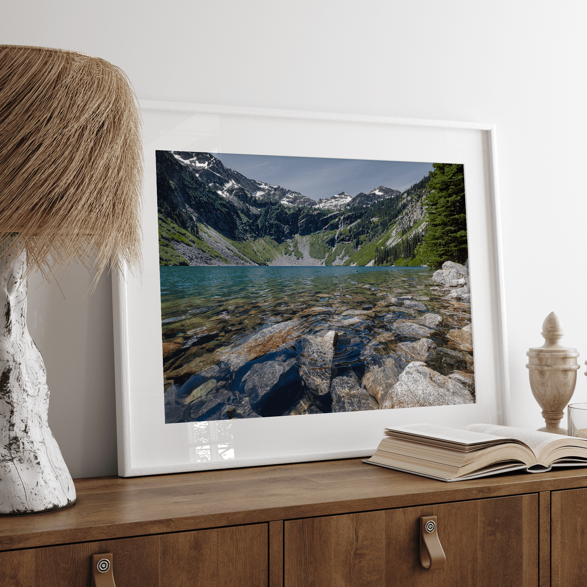 A framed fine art landscape print of a crystal-clear blue lake in Washington State. The tranquil water reveals colorful rocks beneath the surface, with snow-capped mountains towering in the distance.