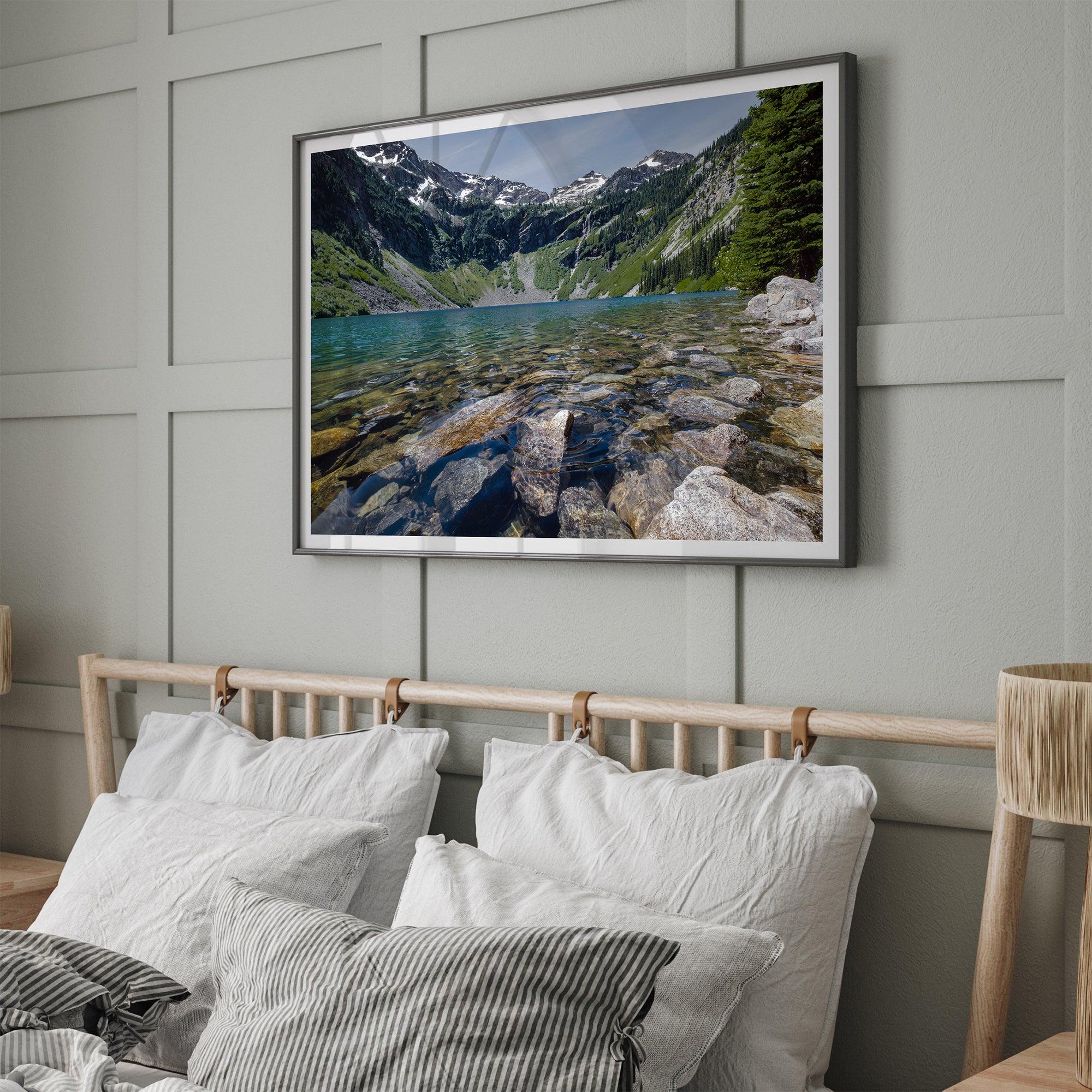 A framed fine art landscape print of a crystal-clear blue lake in Washington State. The tranquil water reveals colorful rocks beneath the surface, with snow-capped mountains towering in the distance.