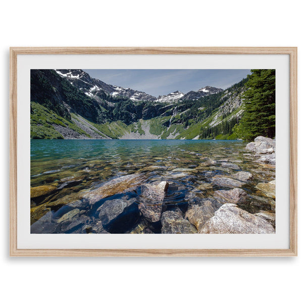 A framed fine art landscape print of a crystal-clear blue lake in Washington State. The tranquil water reveals colorful rocks beneath the surface, with snow-capped mountains towering in the distance.