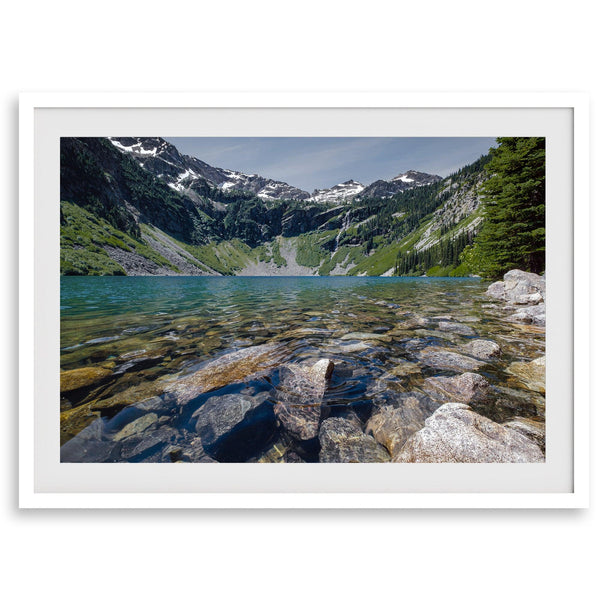 A framed fine art landscape print of a crystal-clear blue lake in Washington State. The tranquil water reveals colorful rocks beneath the surface, with snow-capped mountains towering in the distance.