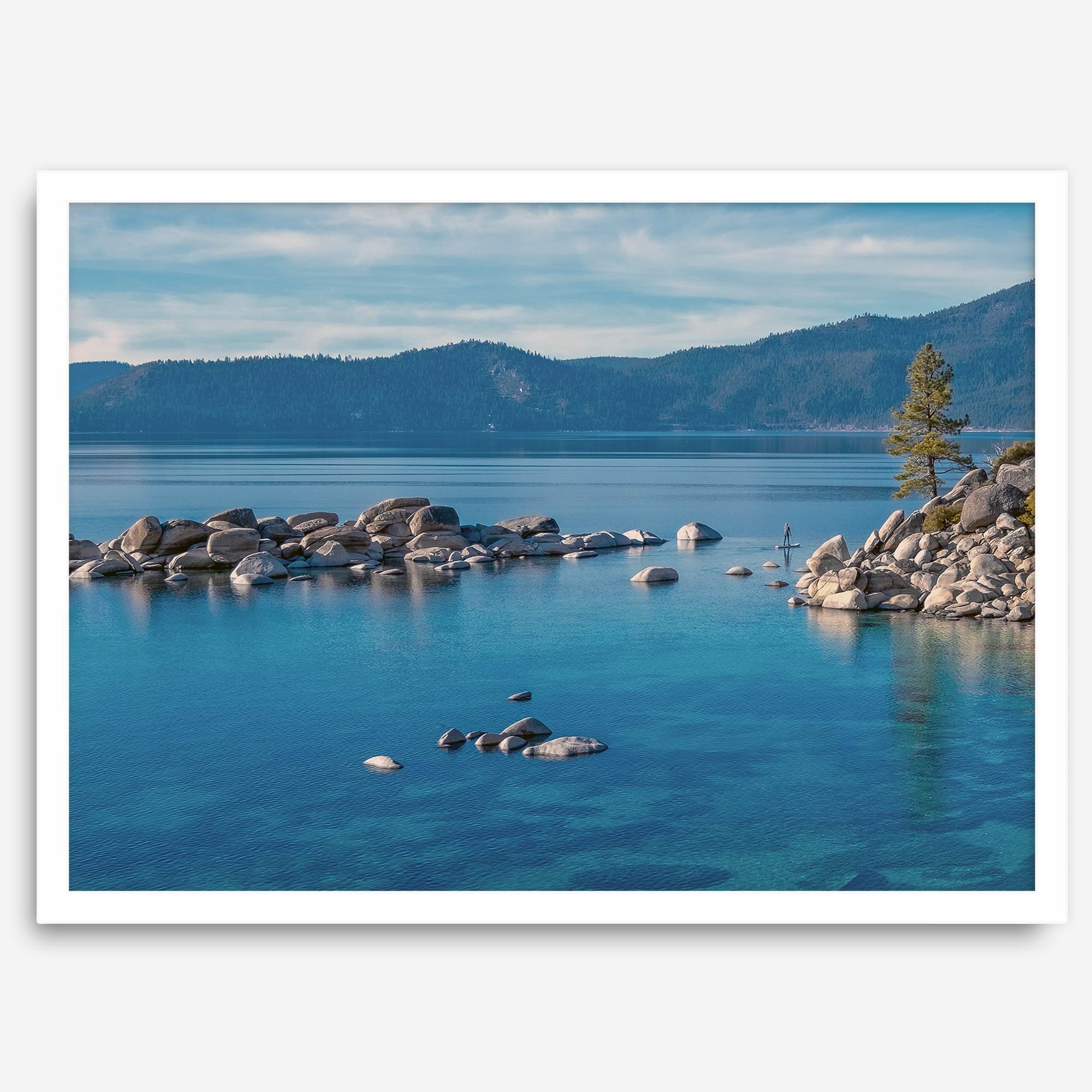 A fine art Lake Tahoe print showcasing a lone stand-up paddle peacefully gliding across the clear water lake near the huge rocks of Sand Harbor Beach with the backdrop of the Sierra Nevada mountains.