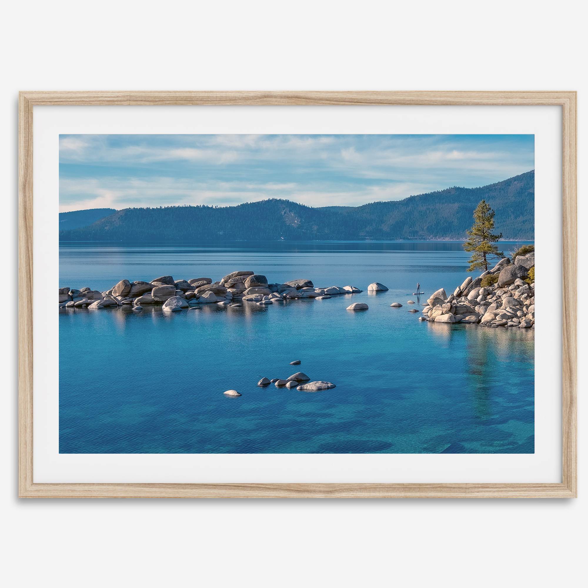 A fine art Lake Tahoe print showcasing a lone stand-up paddle peacefully gliding across the clear water lake near the huge rocks of Sand Harbor Beach with the backdrop of the Sierra Nevada mountains.