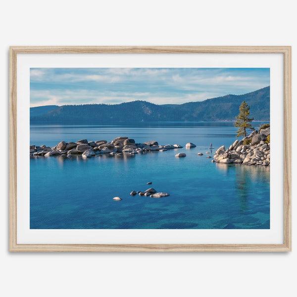 A fine art Lake Tahoe print showcasing a lone stand-up paddle peacefully gliding across the clear water lake near the huge rocks of Sand Harbor Beach with the backdrop of the Sierra Nevada mountains.