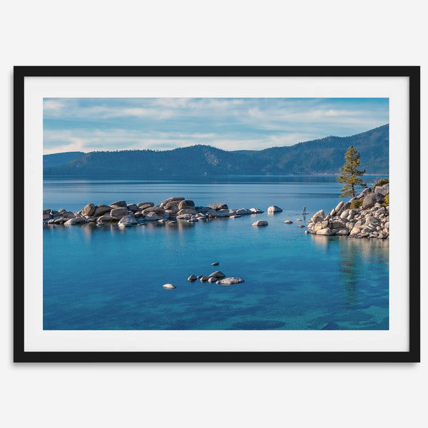 A fine art Lake Tahoe print showcasing a lone stand-up paddle peacefully gliding across the clear water lake near the huge rocks of Sand Harbor Beach with the backdrop of the Sierra Nevada mountains.
