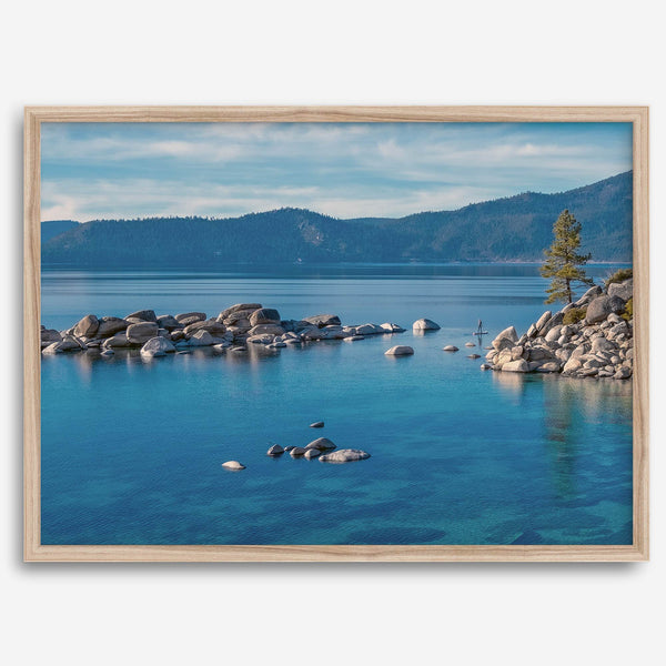 A fine art Lake Tahoe print showcasing a lone stand-up paddle peacefully gliding across the clear water lake near the huge rocks of Sand Harbor Beach with the backdrop of the Sierra Nevada mountains.