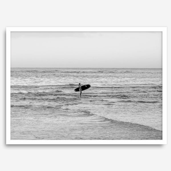 A fine art photography print capturing a female surfer walking back from the ocean at sunset with waves breaking in the foreground, capturing the end of a surf session.