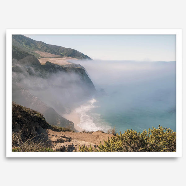 A fine art print of a misty beach in Big Sur, California. Waves crash against the shore as fog drapes over the rugged coastal cliffs, creating a serene, atmospheric scene