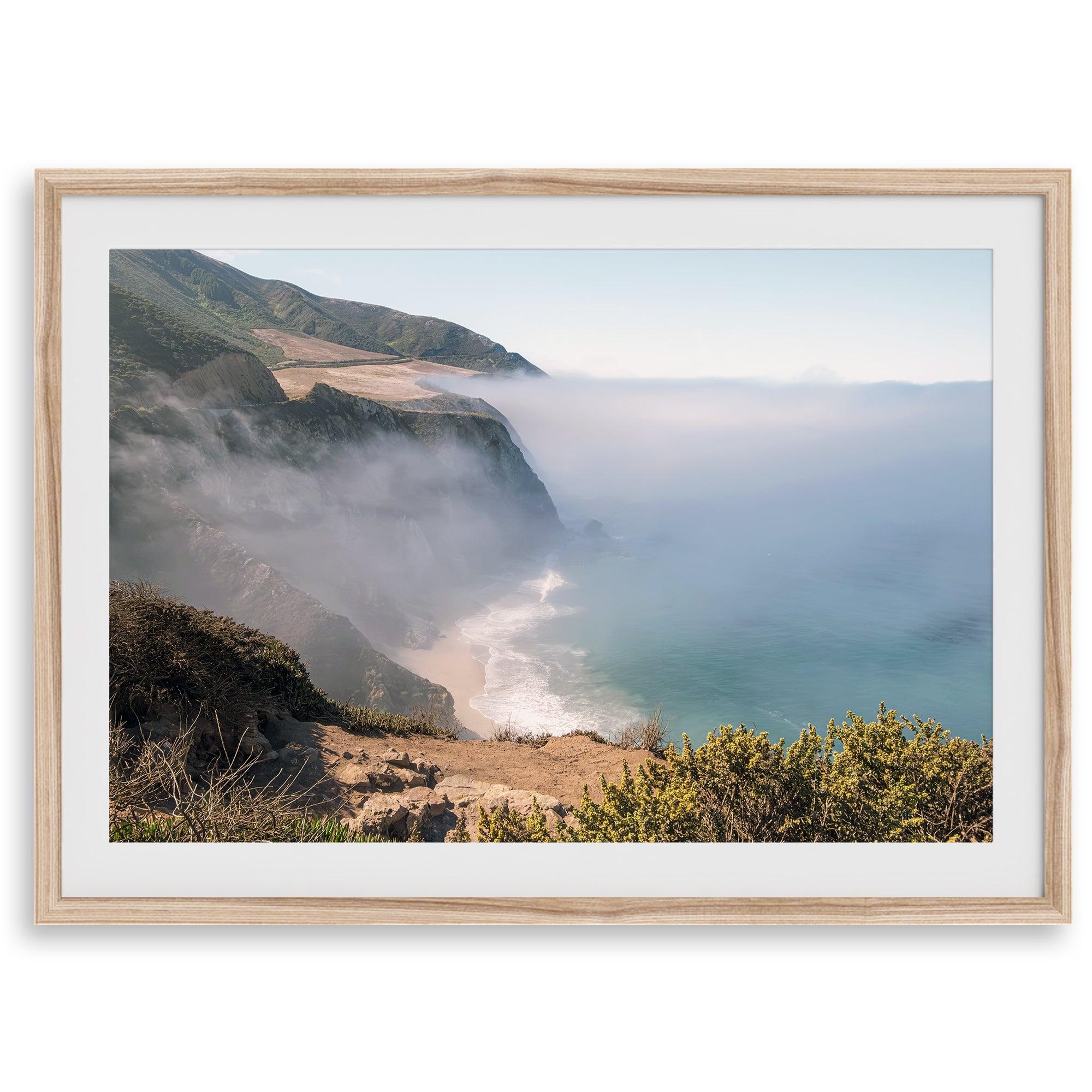 A fine art print of a misty beach in Big Sur, California. Waves crash against the shore as fog drapes over the rugged coastal cliffs, creating a serene, atmospheric scene