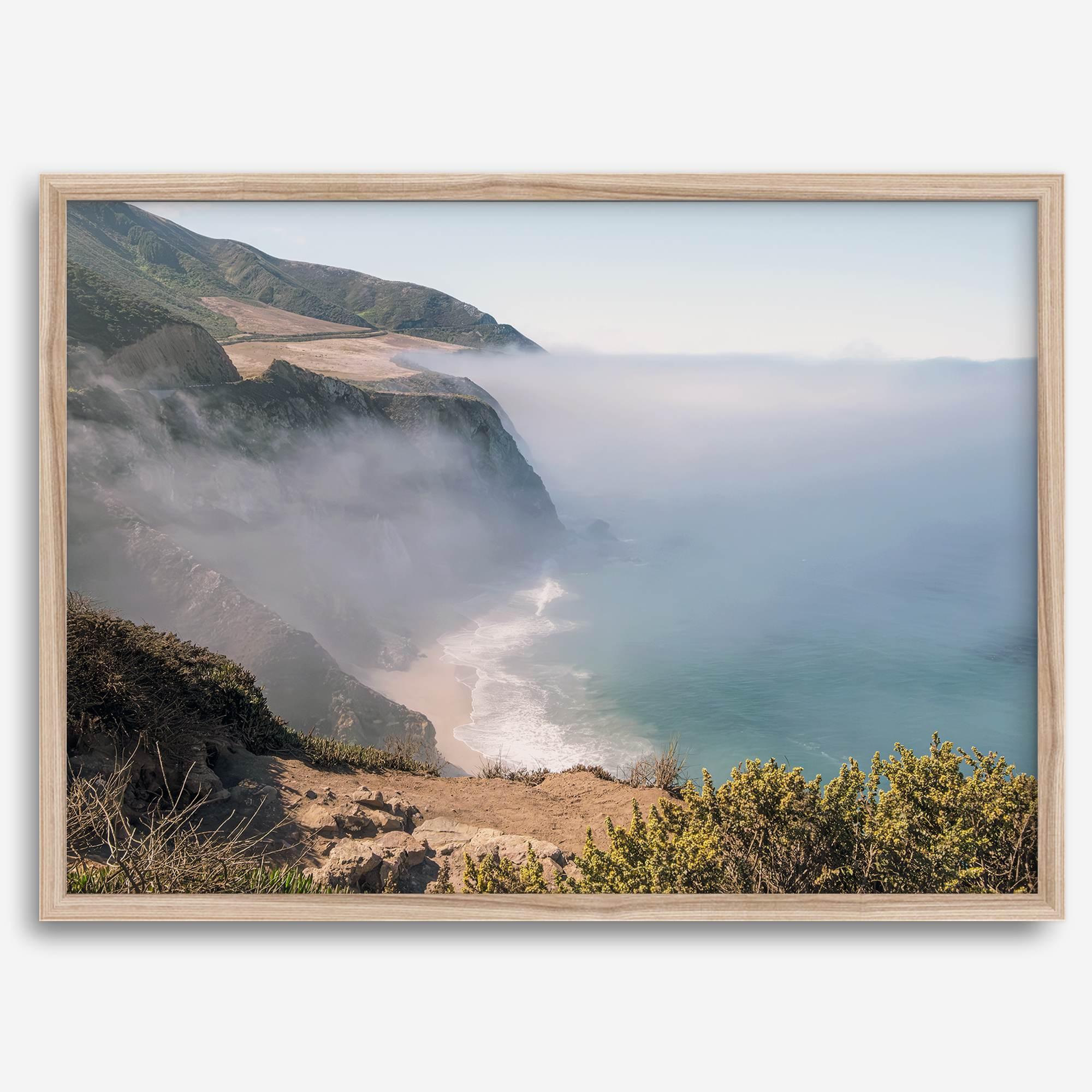 A fine art print of a misty beach in Big Sur, California. Waves crash against the shore as fog drapes over the rugged coastal cliffs, creating a serene, atmospheric scene