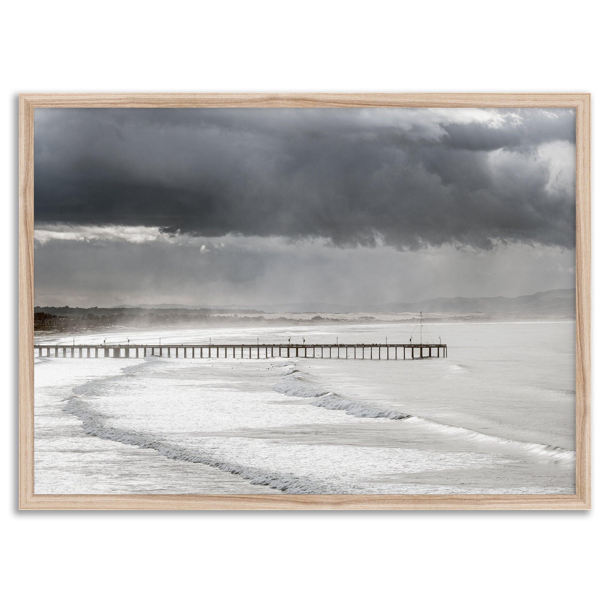 This fine art ocean photography print was taken in Pismo Beach, California, and showcases a distant view of an ocean pier and stormy clouds. Looking long and hard, you can see tens of surfers around the pier.