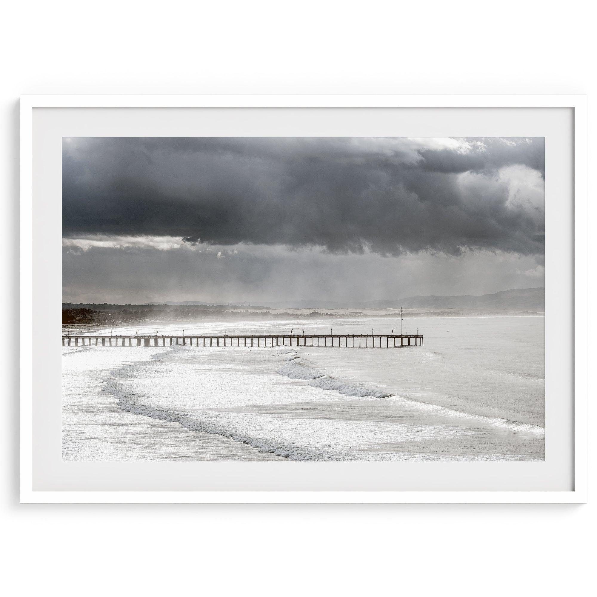 This fine art ocean photography print was taken in Pismo Beach, California, and showcases a distant view of an ocean pier and stormy clouds. Looking long and hard, you can see tens of surfers around the pier.