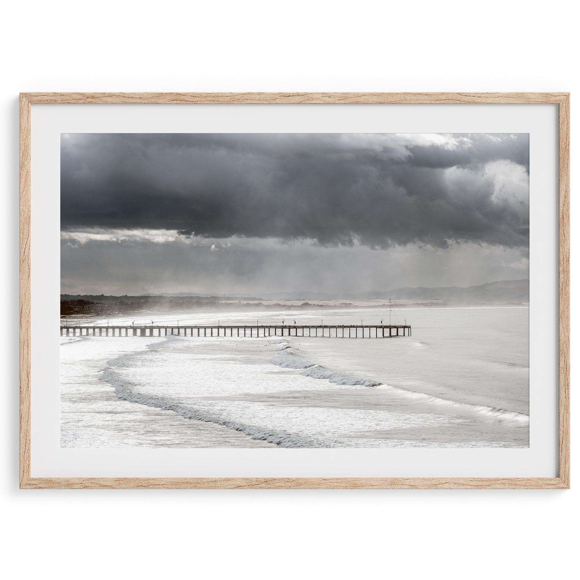 This fine art ocean photography print was taken in Pismo Beach, California, and showcases a distant view of an ocean pier and stormy clouds. Looking long and hard, you can see tens of surfers around the pier.