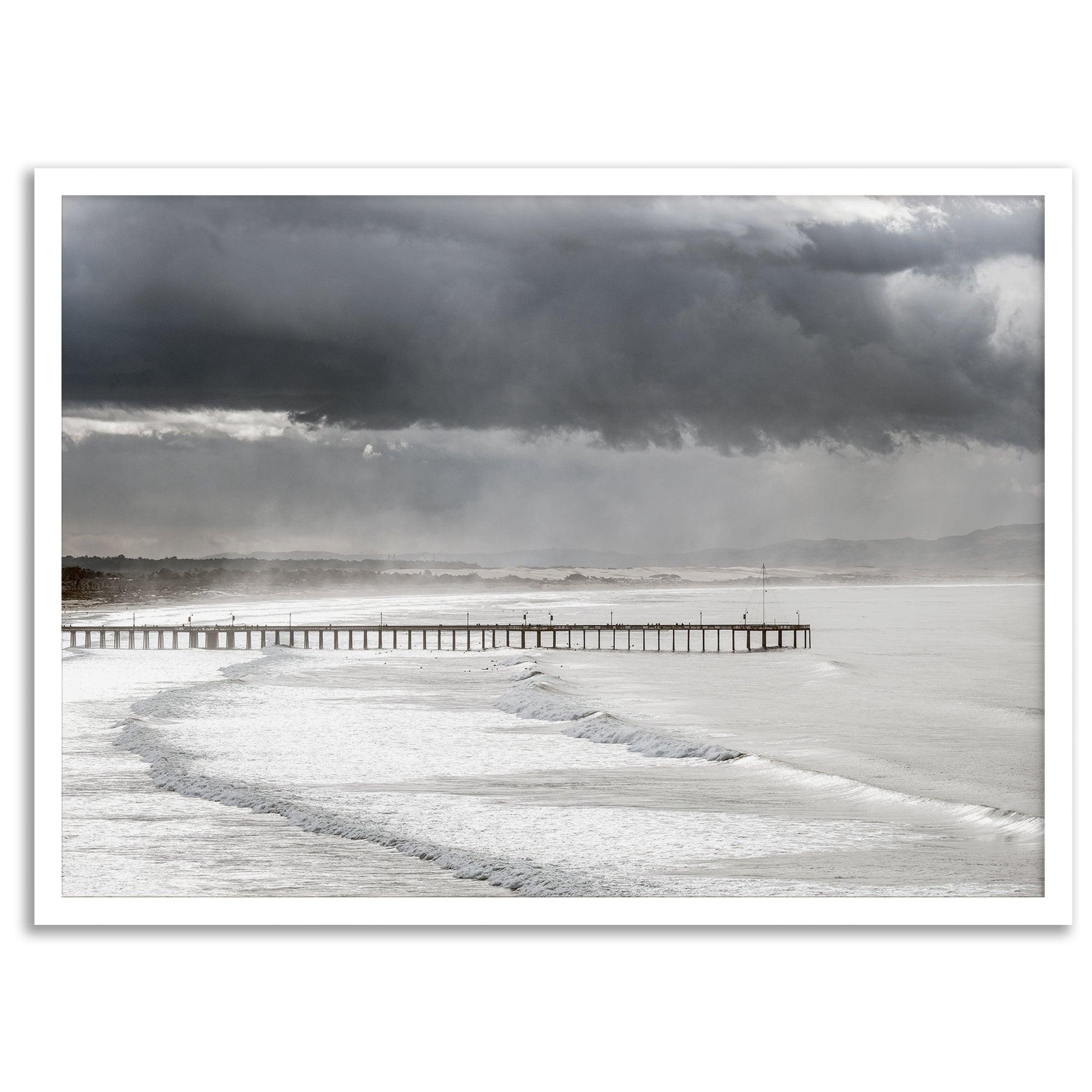 This fine art ocean photography print was taken in Pismo Beach, California, and showcases a distant view of an ocean pier and stormy clouds. Looking long and hard, you can see tens of surfers around the pier.