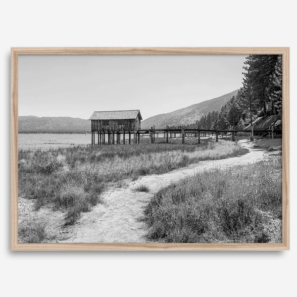 A fine art black and white vintage style framed photography print featuring a rustic old boathouse nested on a Lake Tahoe beach.