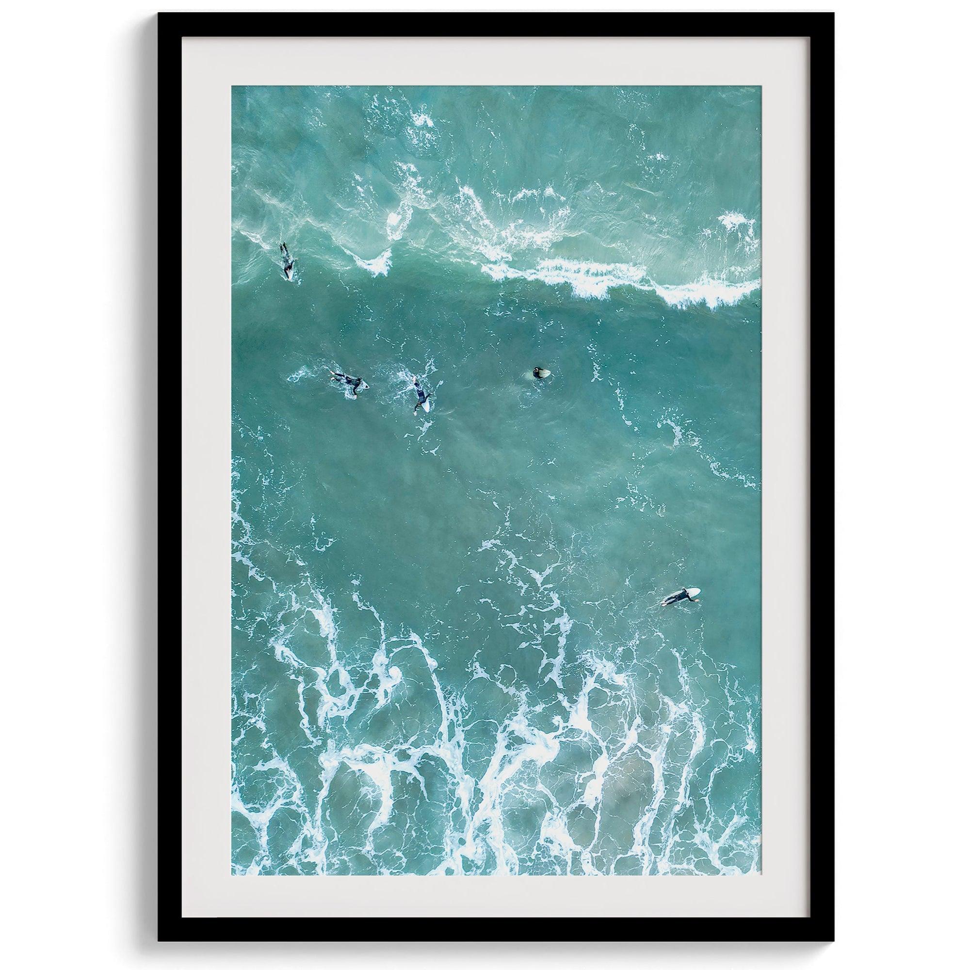 Vertical aerial photo of surfers paddling out to catch a wave at sunset, with wave trails visible in the water.