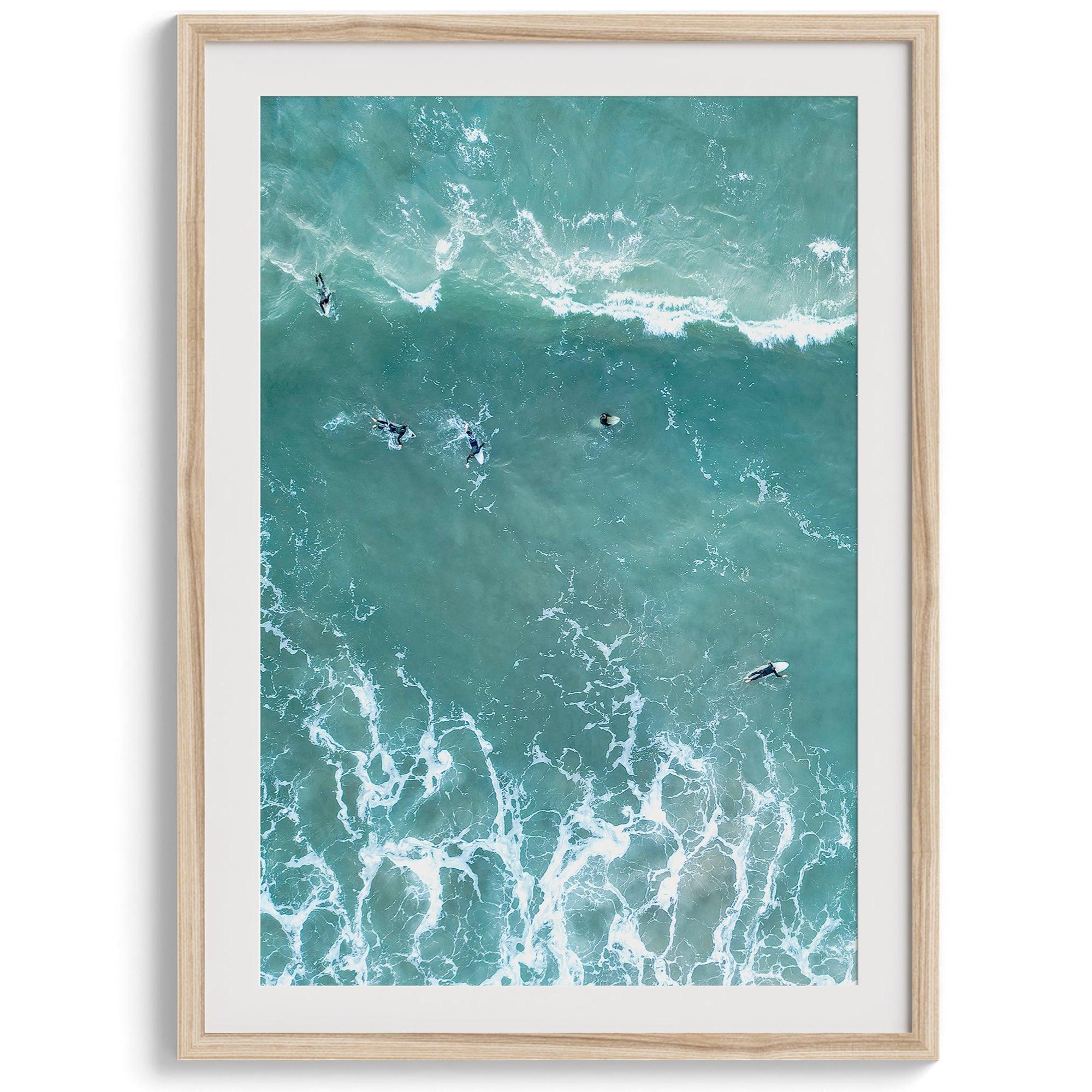 Vertical aerial photo of surfers paddling out to catch a wave at sunset, with wave trails visible in the water.