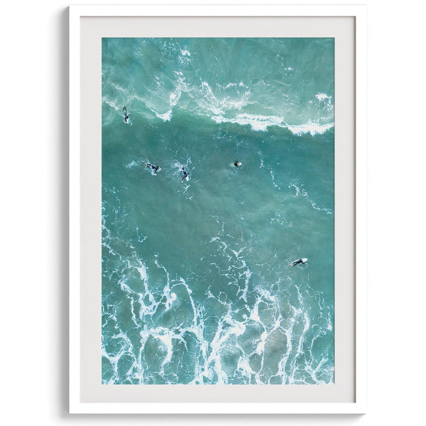 Vertical aerial photo of surfers paddling out to catch a wave at sunset, with wave trails visible in the water.