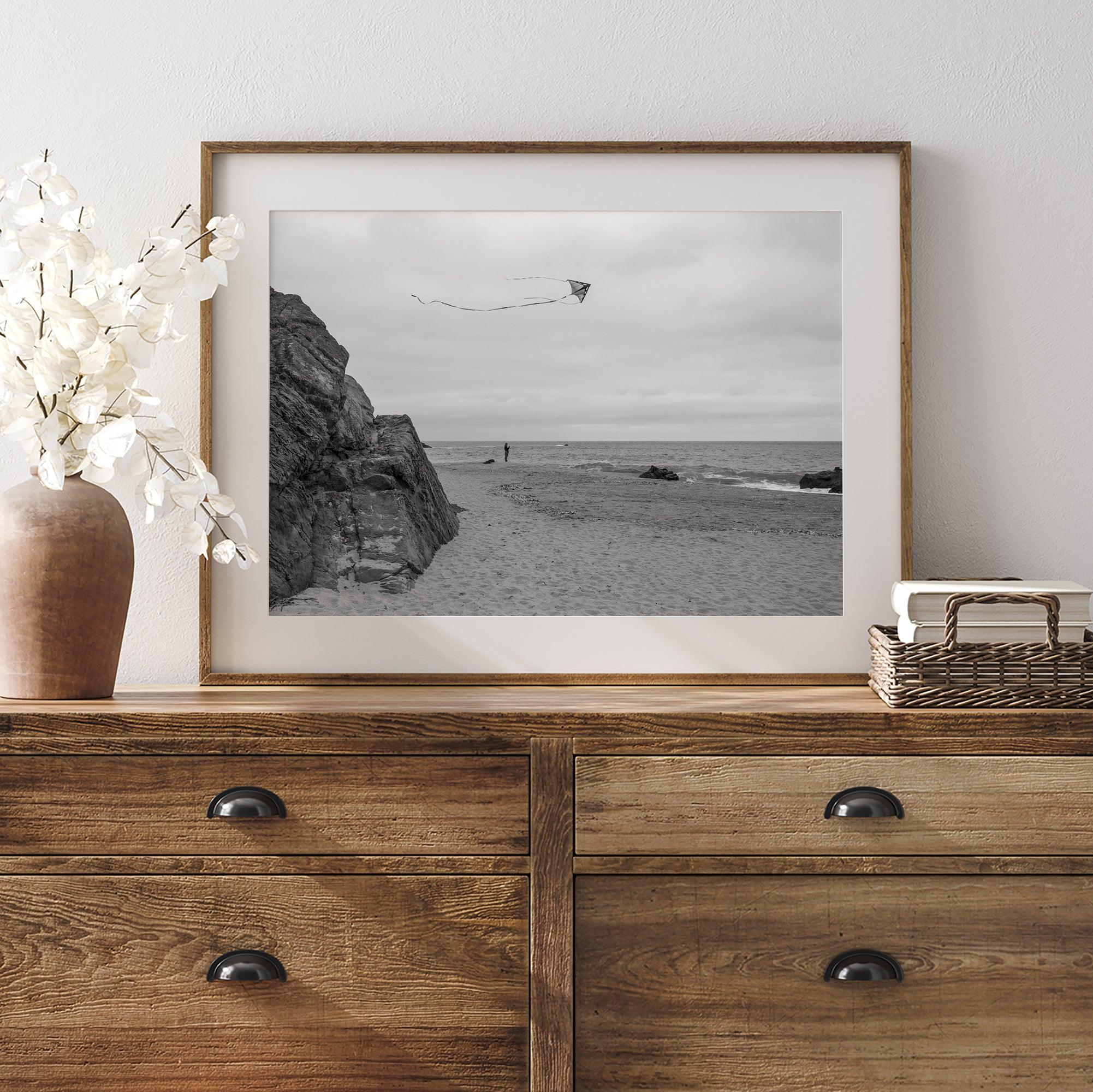 Black-and-white framed photo of a kite flying over a quiet California beach with rocky cliffs and an overcast sky.