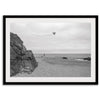 Black-and-white framed photo of a kite flying over a quiet California beach with rocky cliffs and an overcast sky.