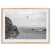 Black-and-white framed photo of a kite flying over a quiet California beach with rocky cliffs and an overcast sky.