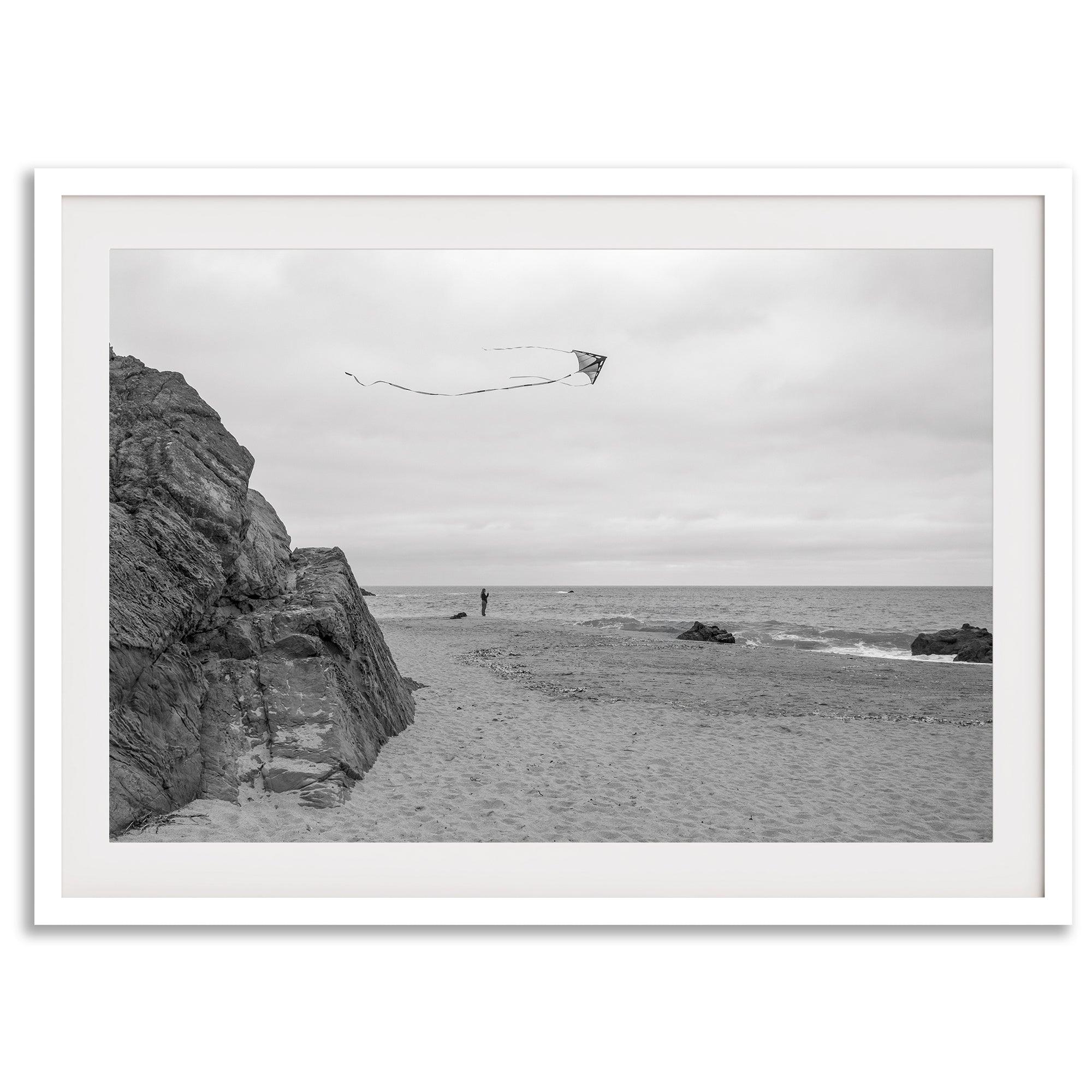 Black-and-white framed photo of a kite flying over a quiet California beach with rocky cliffs and an overcast sky.