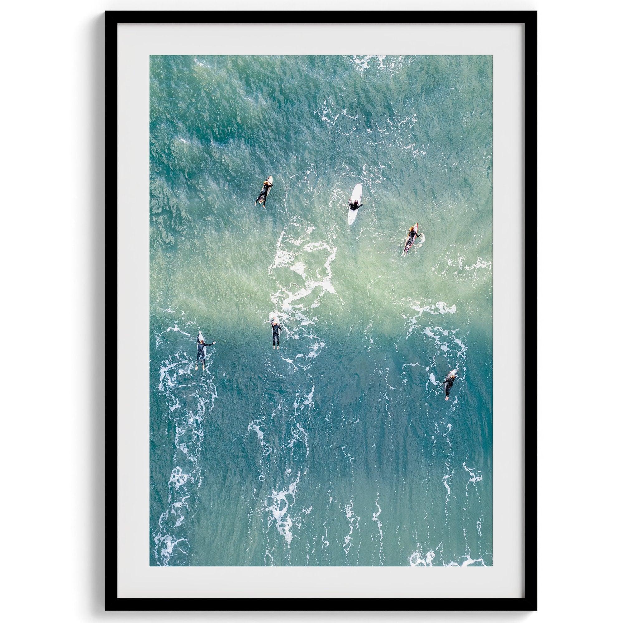 Vertical aerial photo of surfers paddling out to catch a wave at sunset, with wave trails visible in the water.