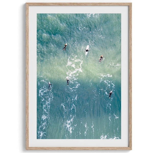 Vertical aerial photo of surfers paddling out to catch a wave at sunset, with wave trails visible in the water.