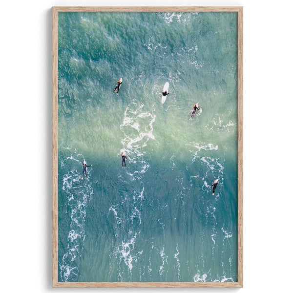 Vertical aerial photo of surfers paddling out to catch a wave at sunset, with wave trails visible in the water.