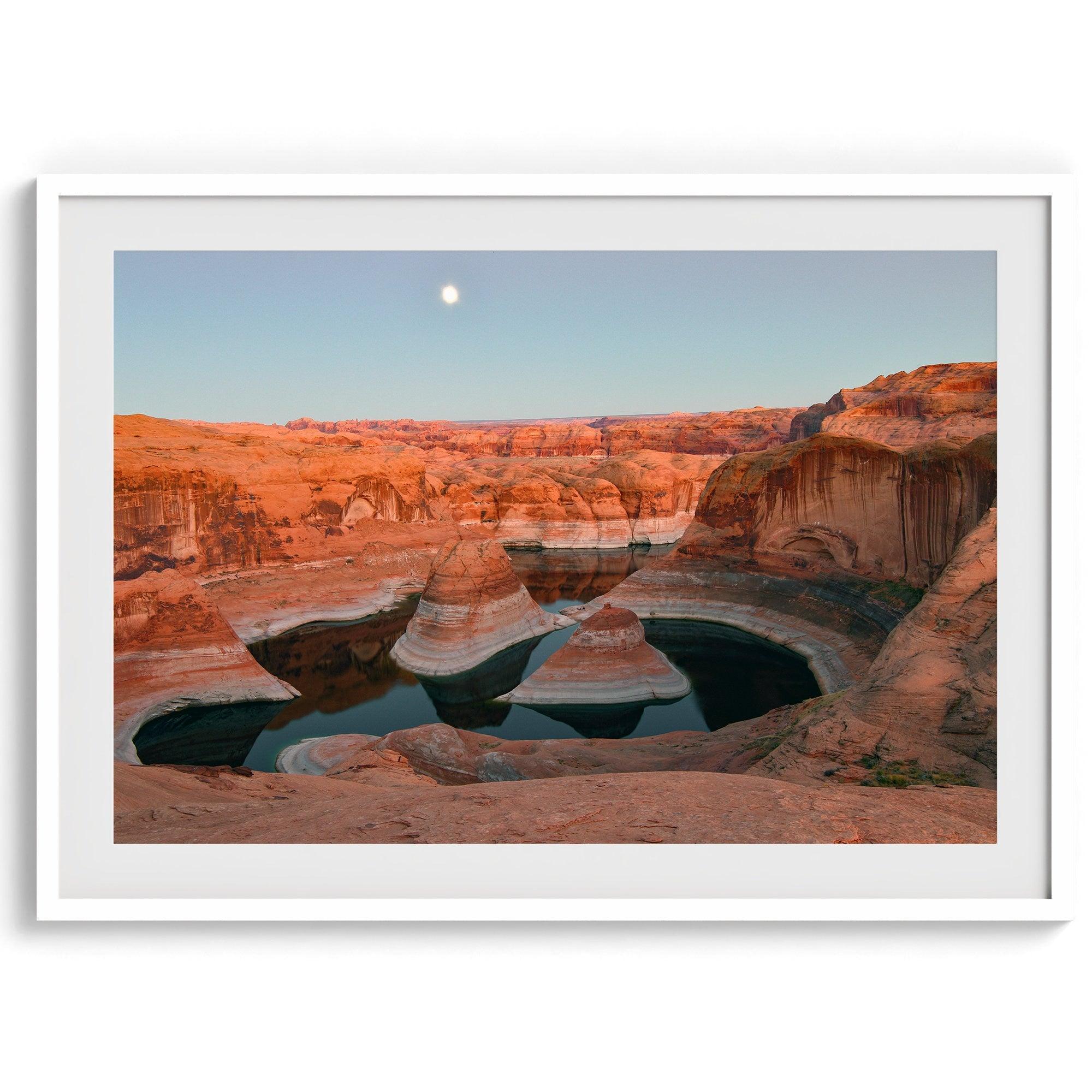 Landscape photo of Reflection Canyon in the Utah desert at sunset, with fiery red hues reflected in still water below a blue sky with a moon.